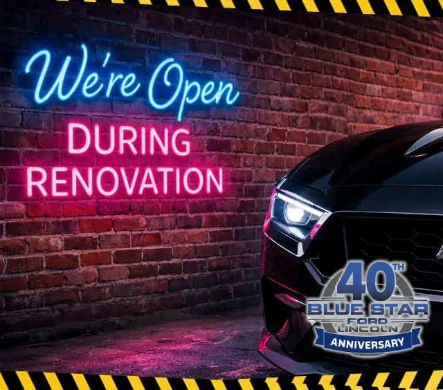 Neon sign on brick wall reads "We're Open During Renovation" beside a car and "40th Blue Star Ford Lincoln Anniversary" badge.
