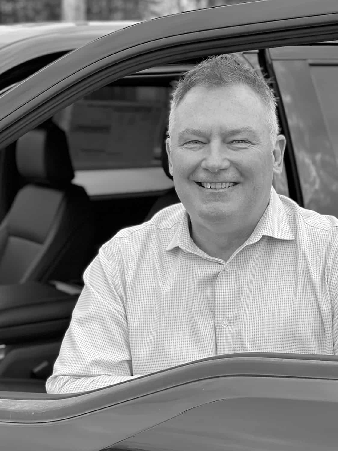 Smiling man sitting in a car driver's seat, viewed through the open window, wearing a checkered shirt.