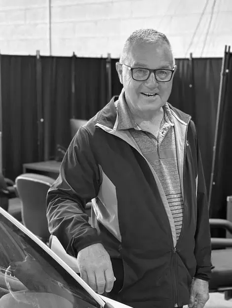 Older man with glasses and a jacket smiling indoors, standing by chairs and a car windshield.