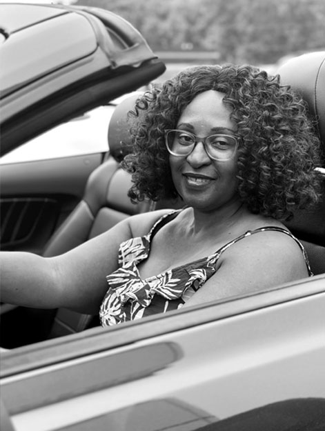 Woman smiling while sitting in a convertible car, wearing glasses and a patterned dress.